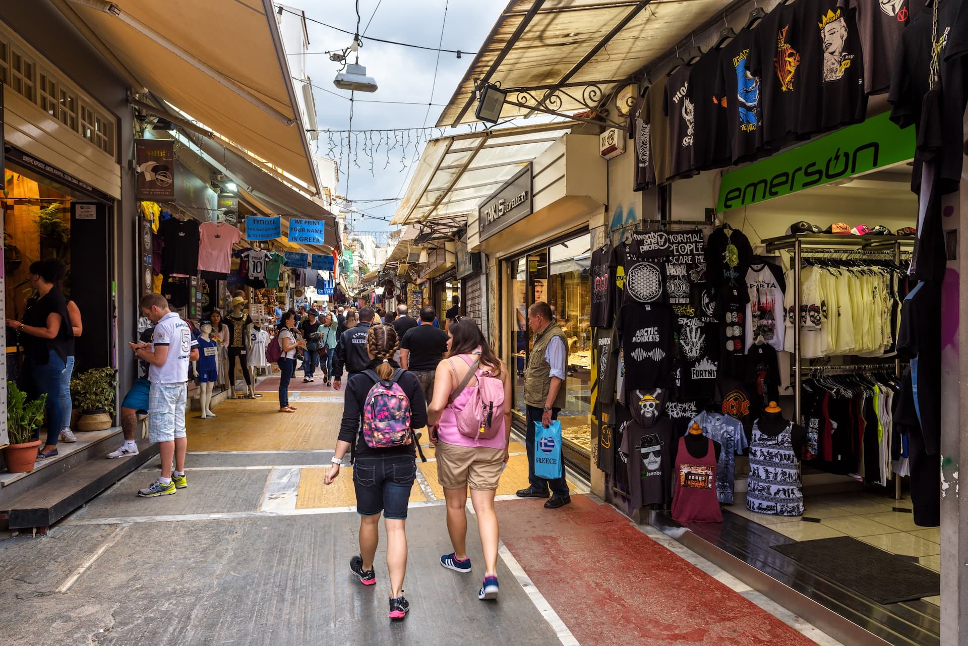 Two people walking through a market street lined with shops and stalls selling various items. - Olive Oil Times