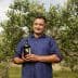 Man standing in an olive grove holding a bottle of olive oil with olive trees in the background. - Olive Oil Times