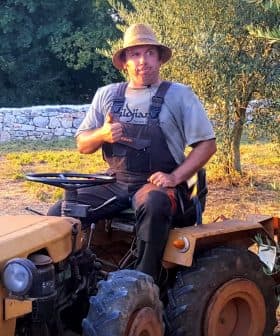 A man sitting on a yellow tractor wearing a straw hat and overalls, giving a thumbs up. - Olive Oil Times