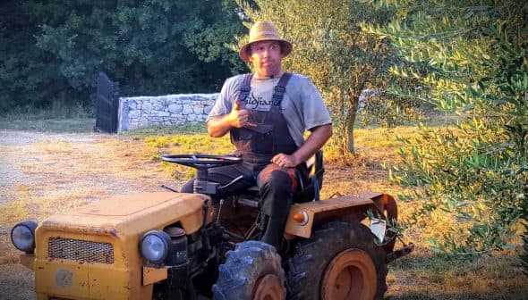 A man sitting on a yellow tractor wearing a straw hat and overalls, giving a thumbs up. - Olive Oil Times