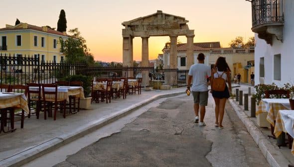 A couple walking along a street near ancient ruins at sunset, with outdoor dining tables visible. - Olive Oil Times