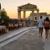 A couple walking along a street near ancient ruins at sunset, with outdoor dining tables visible. - Olive Oil Times