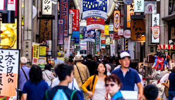 Crowd of people walking through a bustling street market filled with colorful signs in Japan. - Olive Oil Times