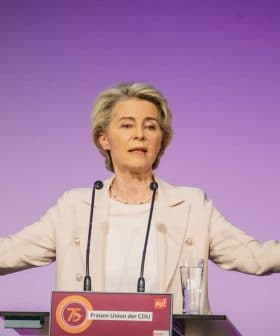 A woman gesturing while speaking at a podium during an event with a purple background. - Olive Oil Times