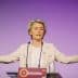 A woman gesturing while speaking at a podium during an event with a purple background. - Olive Oil Times