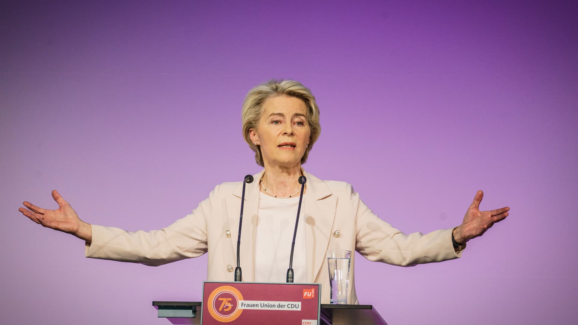 A woman gesturing while speaking at a podium during an event with a purple background. - Olive Oil Times