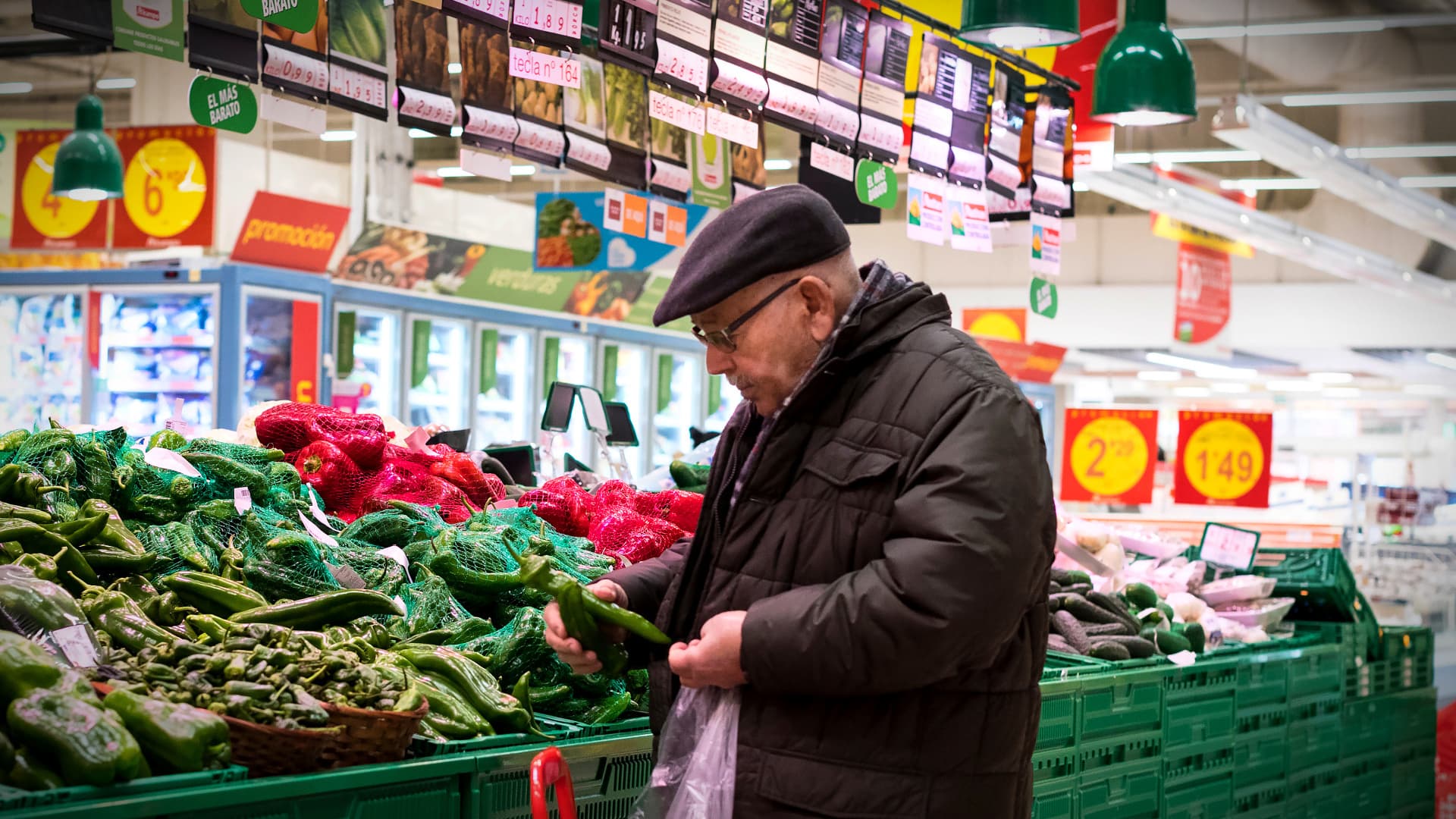 An elderly man examining vegetables in a grocery store with green produce bins. - Olive Oil Times