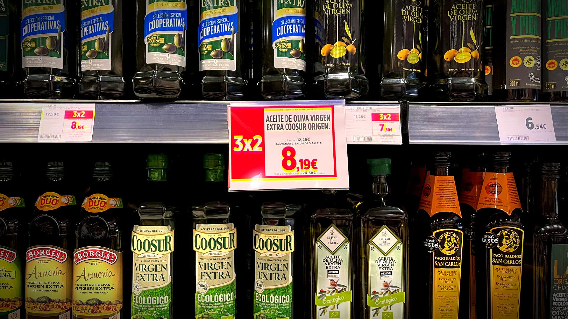 Various bottles of olive oil displayed on a supermarket shelf with pricing labels. - Olive Oil Times