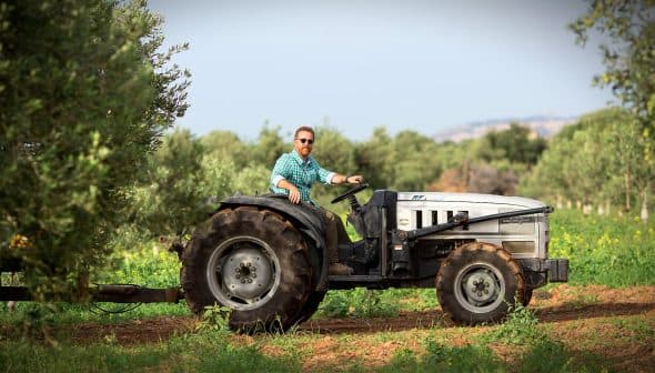 A man with a beard driving a tractor through an olive grove with trees in the background. - Olive Oil Times