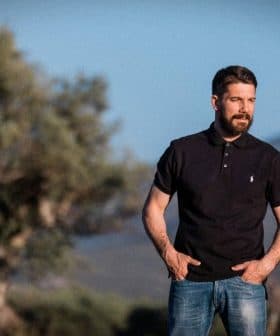 Man wearing a black polo shirt and jeans standing outdoors with olive trees in the background. - Olive Oil Times