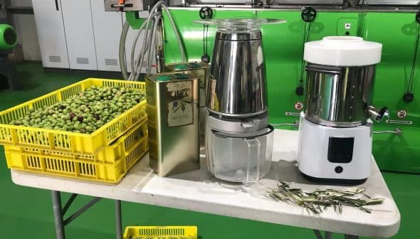Table displaying olive oil extraction equipment alongside baskets of olives. - Olive Oil Times