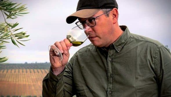 Man in a green shirt and cap tasting olive oil from a glass in a vineyard setting. - Olive Oil Times