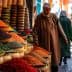 A man in traditional attire walking past colorful spice displays in a market setting. - Olive Oil Times