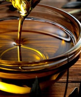 Golden olive oil being poured into a glass bowl placed on a wooden surface. - Olive Oil Times
