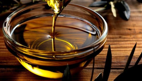 Golden olive oil being poured into a glass bowl placed on a wooden surface. - Olive Oil Times