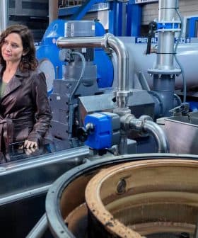 A woman in a brown leather jacket conversing with a man in a factory filled with machinery and equipment. - Olive Oil Times