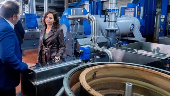 A woman in a brown leather jacket conversing with a man in a factory filled with machinery and equipment. - Olive Oil Times