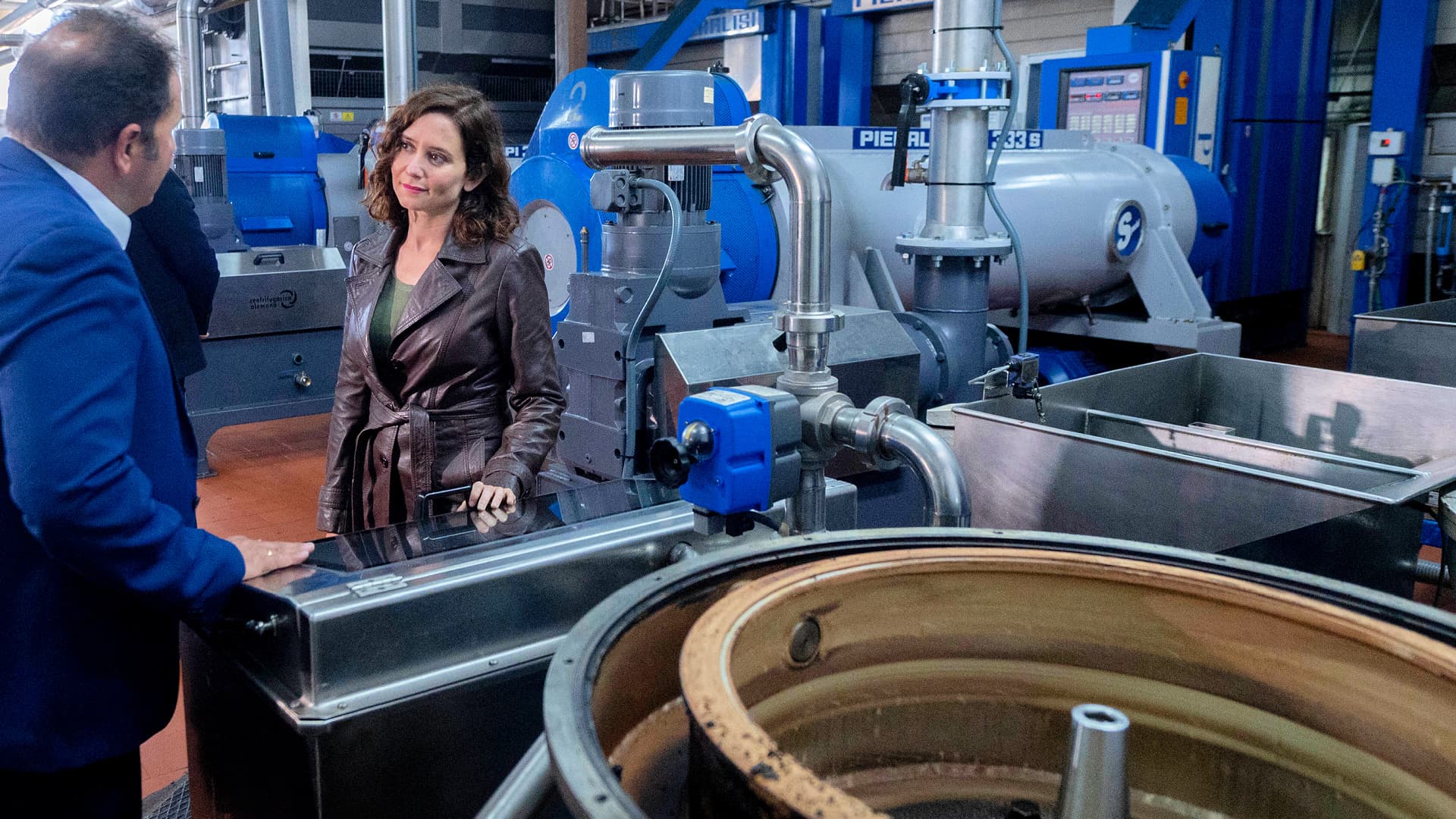 A woman in a brown leather jacket conversing with a man in a factory filled with machinery and equipment. - Olive Oil Times