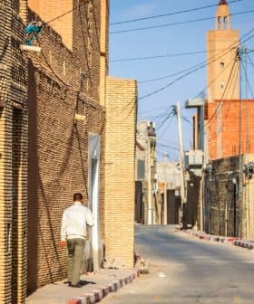 Narrow street lined with brick buildings in a village, with a person walking along the side. - Olive Oil Times