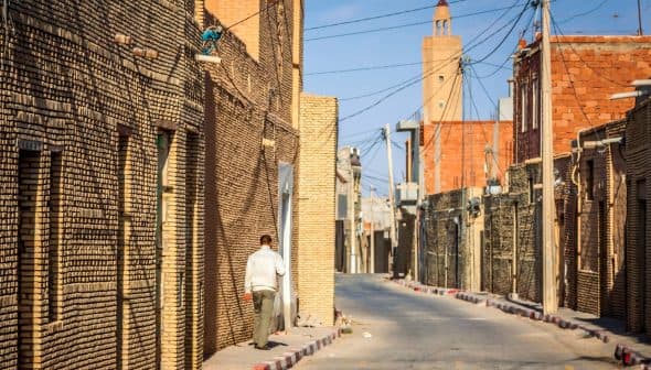 Narrow street lined with brick buildings in a village, with a person walking along the side. - Olive Oil Times