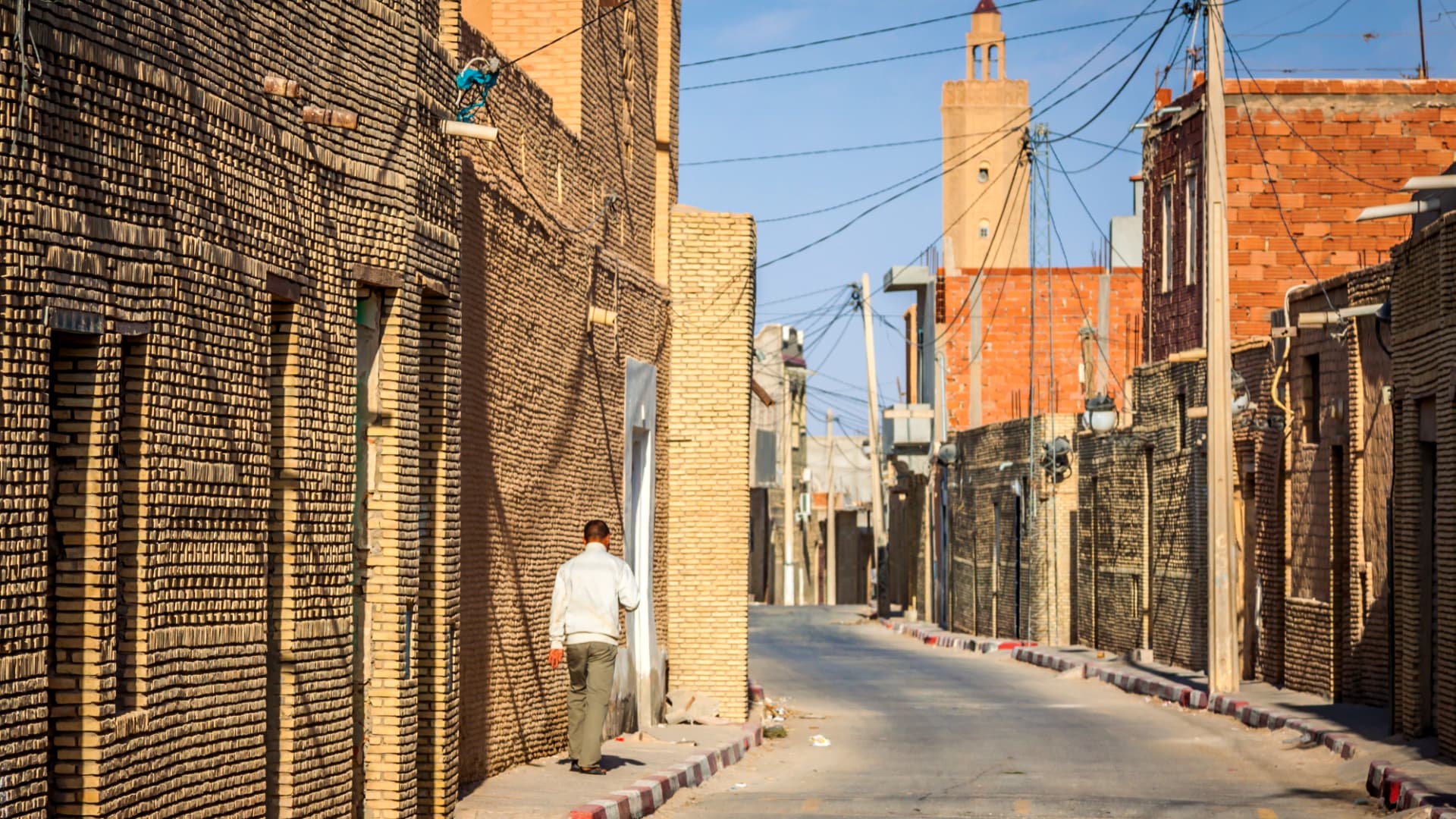 Narrow street lined with brick buildings in a village, with a person walking along the side. - Olive Oil Times