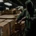 A warehouse worker in a green hoodie sorting through cardboard boxes in a dimly lit environment. - Olive Oil Times