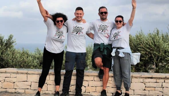 Four individuals posing together outdoors, wearing matching t-shirts and smiling at the camera. - Olive Oil Times