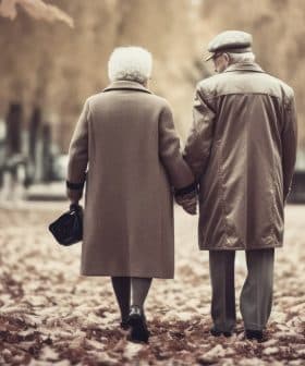 An elderly couple holding hands while walking through a park covered in autumn leaves. - Olive Oil Times
