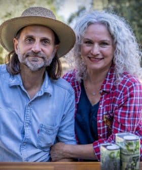 A man and woman sitting together outdoors, both smiling, with cans placed on a table in front of them. - Olive Oil Times