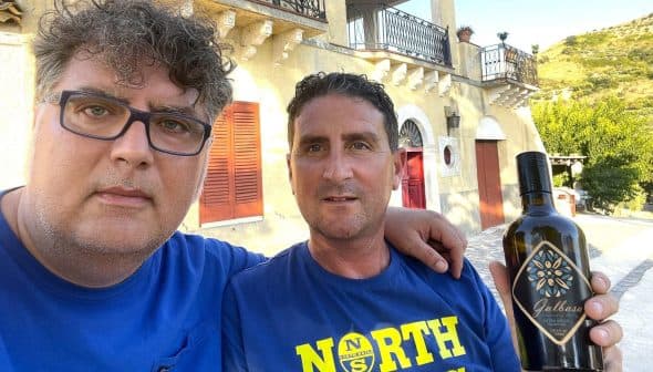 Two men posing for a selfie while holding a bottle of olive oil outdoors. - Olive Oil Times