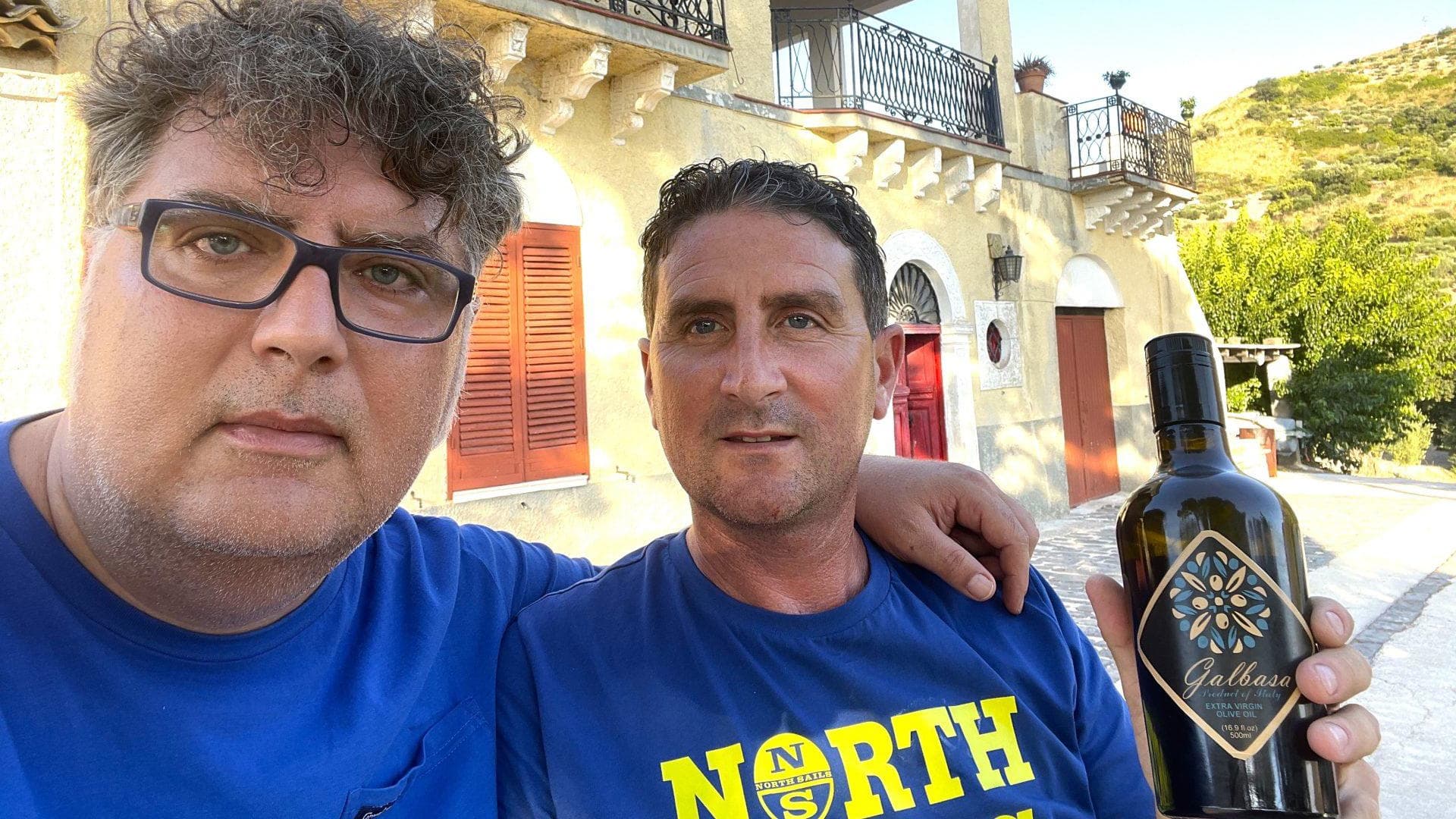 Two men posing for a selfie while holding a bottle of olive oil outdoors. - Olive Oil Times