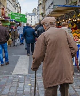 Elderly man with a cane walking through a market street lined with stalls and produce. - Olive Oil Times