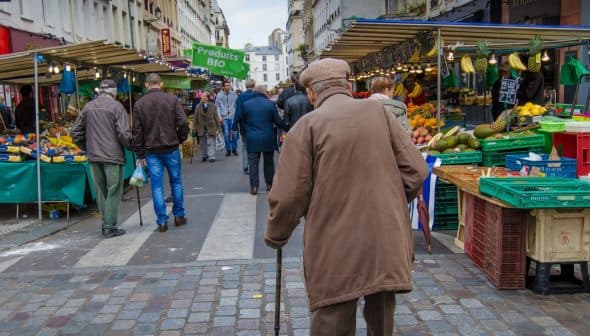 Elderly man with a cane walking through a market street lined with stalls and produce. - Olive Oil Times
