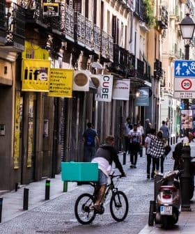 A street scene featuring a bicyclist with a teal delivery box and pedestrians walking in a narrow urban area. - Olive Oil Times