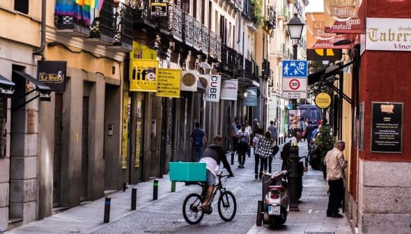 A street scene featuring a bicyclist with a teal delivery box and pedestrians walking in a narrow urban area. - Olive Oil Times