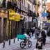 A street scene featuring a bicyclist with a teal delivery box and pedestrians walking in a narrow urban area. - Olive Oil Times