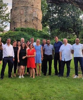 A group of individuals posing together for a photo in an outdoor area with greenery and a stone structure. - Olive Oil Times
