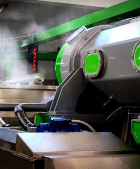 Industrial olive oil processing machine with green accents and steam emerging from the equipment. - Olive Oil Times