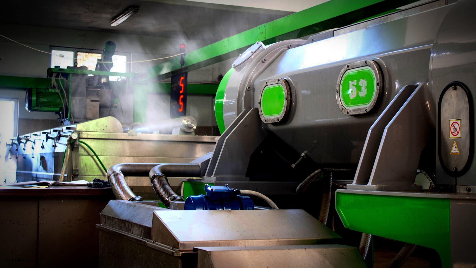 Industrial olive oil processing machine with green accents and steam emerging from the equipment. - Olive Oil Times