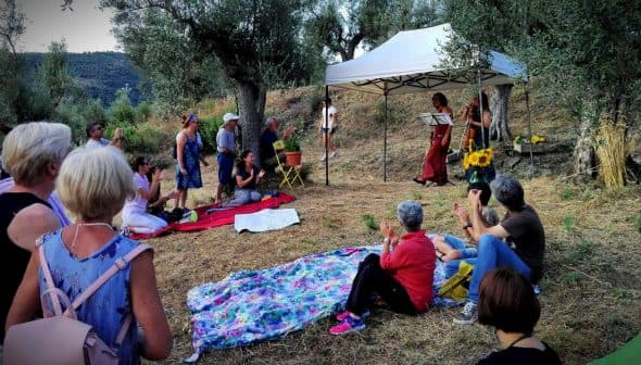 Group of people gathered outdoors under a tent for a performance, with some seated on blankets and others standing. - Olive Oil Times