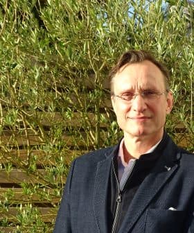 Man wearing glasses and a black jacket standing in front of green plants. - Olive Oil Times