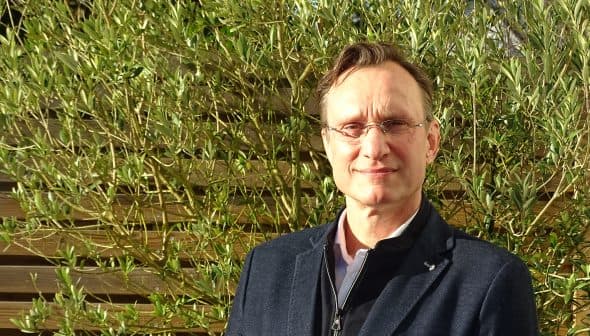 Man wearing glasses and a black jacket standing in front of green plants. - Olive Oil Times