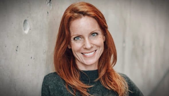 Woman with red hair smiling while leaning against a textured wall. - Olive Oil Times