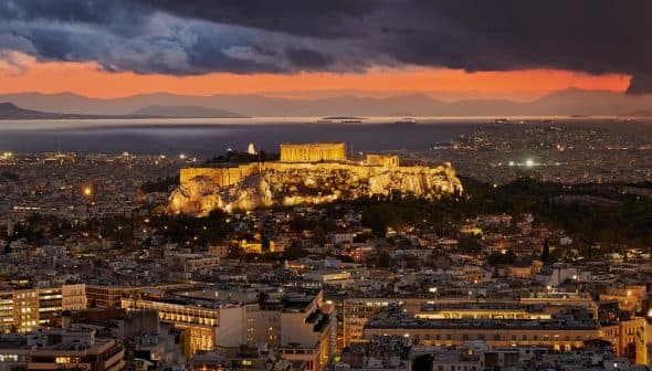 View of the Acropolis of Athens illuminated at sunset with a colorful sky and distant mountains. - Olive Oil Times