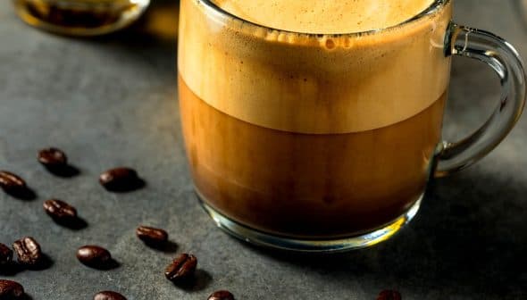 A glass mug filled with a layered coffee beverage, featuring a frothy top and dark liquid below. - Olive Oil Times