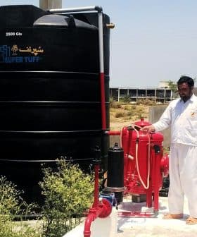 A large black water storage tank labeled 'SUPER TUFF' next to a red pump system and a man in white clothing. - Olive Oil Times