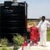 A large black water storage tank labeled 'SUPER TUFF' next to a red pump system and a man in white clothing. - Olive Oil Times