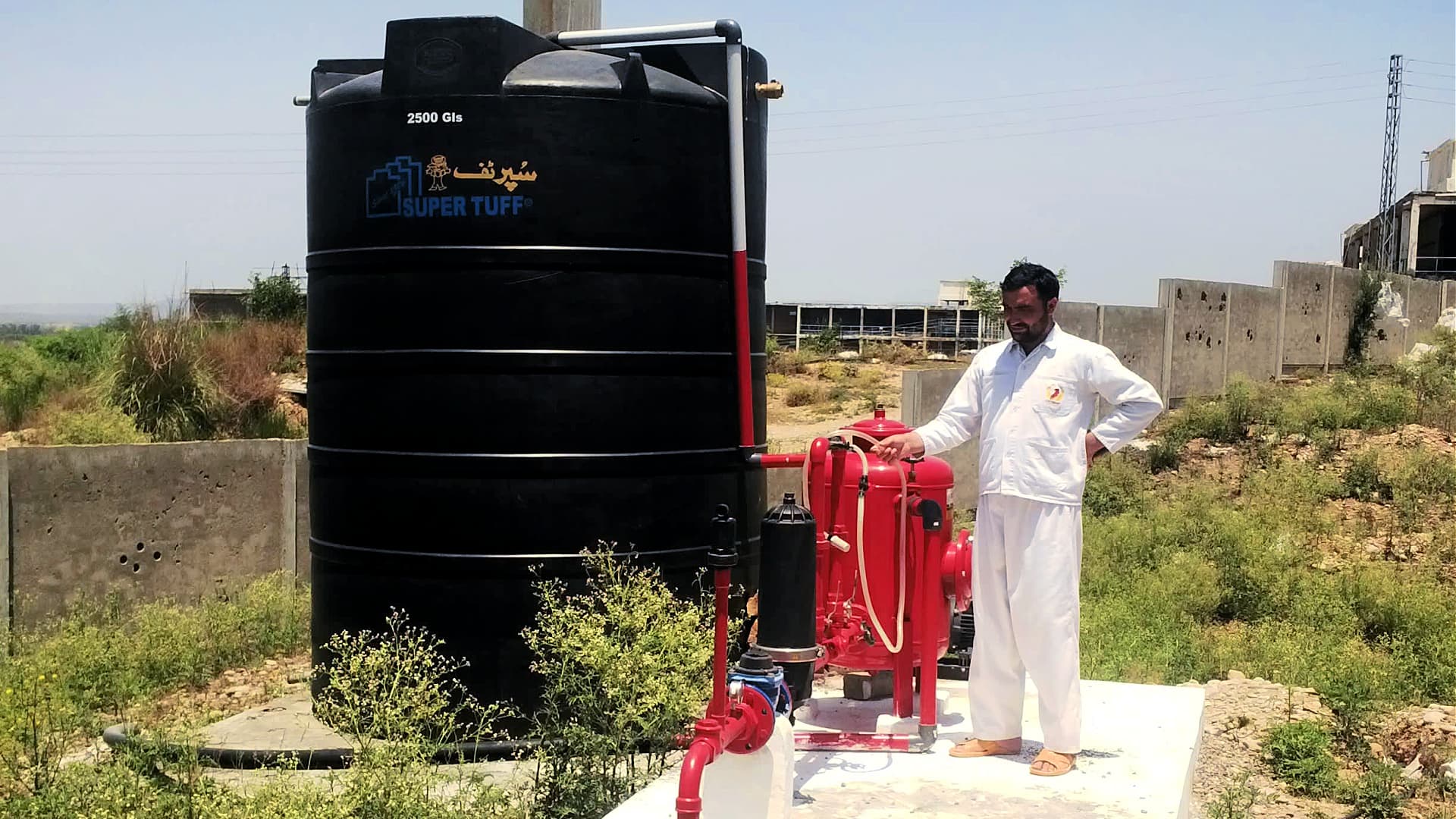 A large black water storage tank labeled 'SUPER TUFF' next to a red pump system and a man in white clothing. - Olive Oil Times