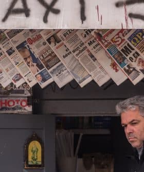 A man standing in front of a newsstand displaying various newspapers and magazines. - Olive Oil Times
