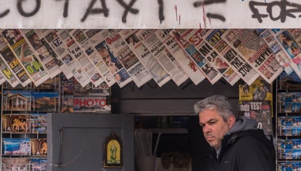 A man standing in front of a newsstand displaying various newspapers and magazines. - Olive Oil Times